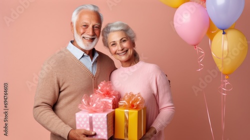 Senior couple smiling with a birthday gift and pastel balloons in a studio with a pink background.