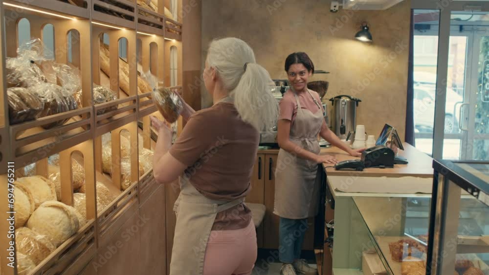 Senior female worker putting bread on shelf and then helping young colleague at cash register in bakery Senior female worker putting bread on shelf and then helping young colleague at cash register in bakery