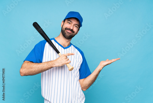 Young man playing baseball over isolated blue background holding copyspace imaginary on the palm to insert an ad