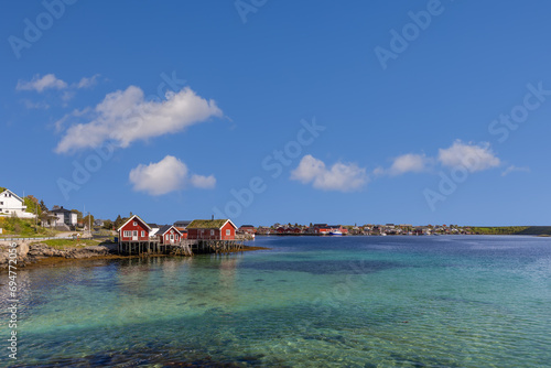 Emerald waters of the Norwegian Sea lap against the red rorbu of Reine, Lofoten Islands, under a sky dotted with fluffy clouds photo