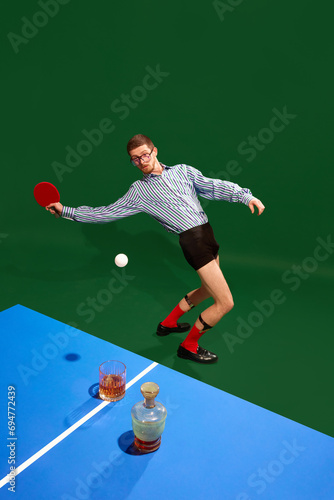 Top view. Funny creative photo with man un shirt and underweared playing table tennis with whiskey glass over green background. Concept of sport, leisure, hobby, creativity, fun and joy. Pop art