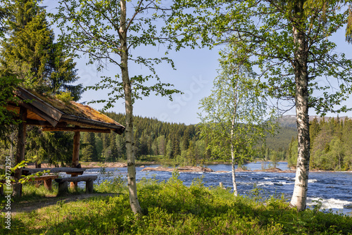 A picnic spot by the Namsen River, Norway, with a mossy-roofed shelter and wooden benches, offering a view of flowing waters amid the lush Trondelag region photo