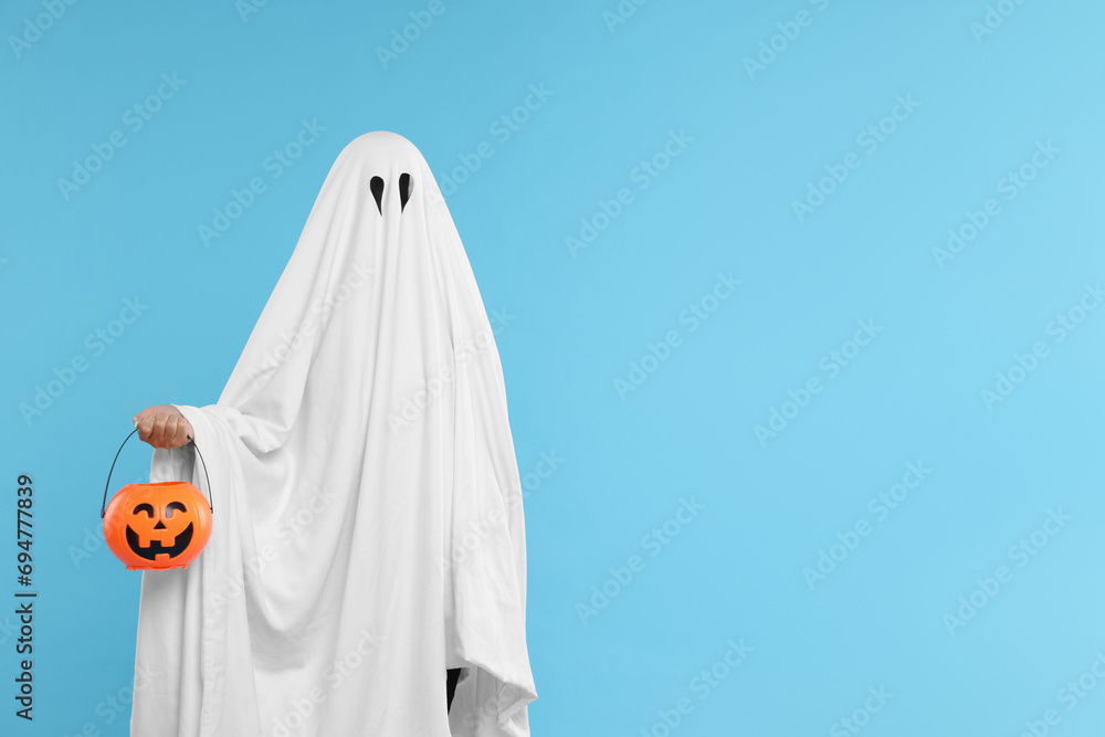 Woman in white ghost costume holding pumpkin bucket on light blue background, space for text. Halloween celebration