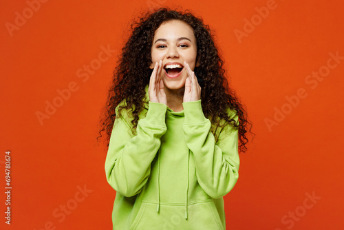 Young promoter fun woman of African American ethnicity she wear green hoody casual clothes scream sharing hot news about sales discount with hands near mouth isolated on plain red orange background.