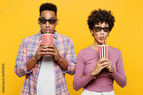 Young amazed surprised couple two friend family man woman wear purple casual clothes wear 3d glasses together watch movie film hold cup of soda pop in cinema look camera isolated on yellow background