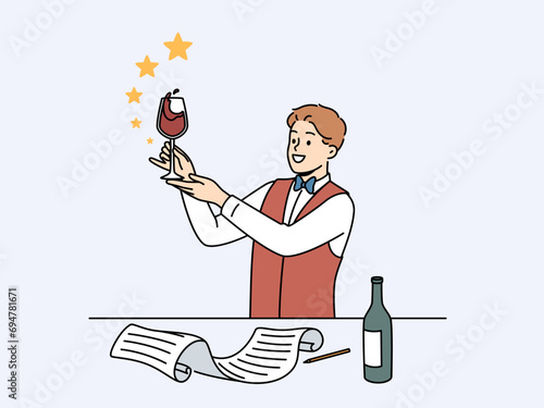 Man sommelier holds glass red wine and evaluates taste of noble alcoholic drink standing near table with bottle and sheet of paper. Guy studying at sommelier school wanting to get job in restaurant