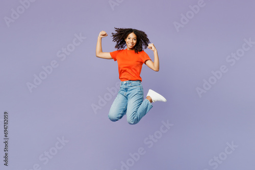 Full body little kid teen girl of African American ethnicity wears orange t-shirt jump high do winner gesture clench fist isolated on plain pastel purple background studio Childhood lifestyle concept