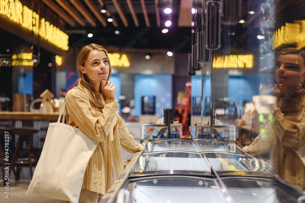 Young customer thoughtful woman wear casual clothes bag standing near refrigerator shopping at supermaket store grocery shop buy choose products inside hypermarket. Purchasing gastronomy food concept. Young customer thoughtful woman wear casual clothes bag standing near refrigerator shopping at supermaket store grocery shop buy choose products inside hypermarket. Purchasing gastronomy food concept.
