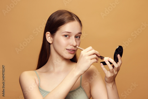 Portrait of attractive young woman with long brunette hair looks at camera and holds cosmetic product against beige background. photo