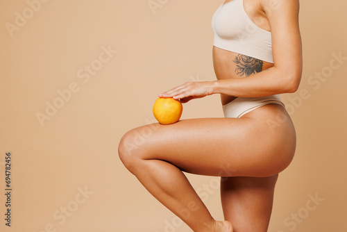 Side view close up cropped young nice lady woman with slim body perfect skin wear nude top bra lingerie stand hold orange on hip isolated on plain pastel beige background. Lifestyle diet fit concept.