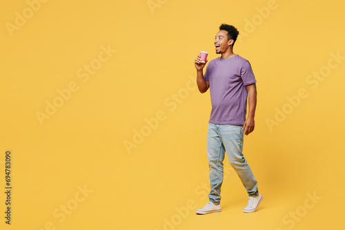 Full body young man of African American ethnicity wears purple t-shirt casual clothes hold takeaway delivery craft paper brown cup coffee to go isolated on plain yellow background. Lifestyle concept.