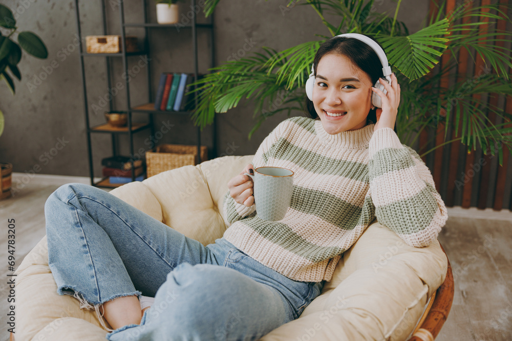 Obraz premium Sideways young woman wears casual clothes listen to music in headphones drink tea sits on armchair stay at home hotel flat rest relax spend free spare time in grey living room indoor. Lounge concept.