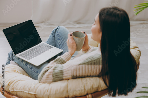 Young IT woman wears casual clothes sits on armchair work use blank screen laptop pc computer drink coffee stay at home hotel flat rest relax spend free spare time in grey living room. Lounge concept.