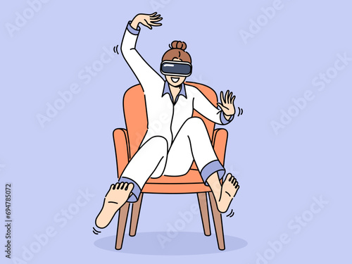 Woman wearing vr headset sits in chair and waves arms and legs, feeling euphoria of being immersed in virtual reality. Girl in pajamas uses vr glasses to travel through metaverse and play video games.