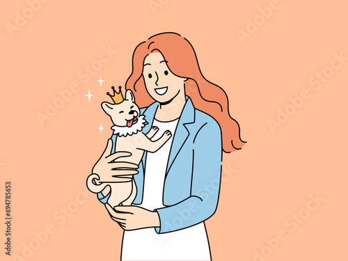 Woman holds small dog wearing crown in arms and smiles enjoying guardianship over puppy. Happy girl loves animals rejoices in presence of purebred dog and looks at screen with smile