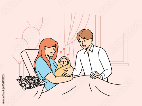 Mother with newborn baby sits on couch in hospital and shows infant to happy father. Smiling married couple have become parents for first time admire sleeping newborn wrapped in blanket