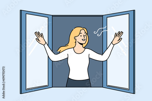 Woman opens window enjoying fresh air coming from street into apartment, and smilingly looks at passers-by. Girl stands near window and wants to check room to get rid of unpleasant smell in house.