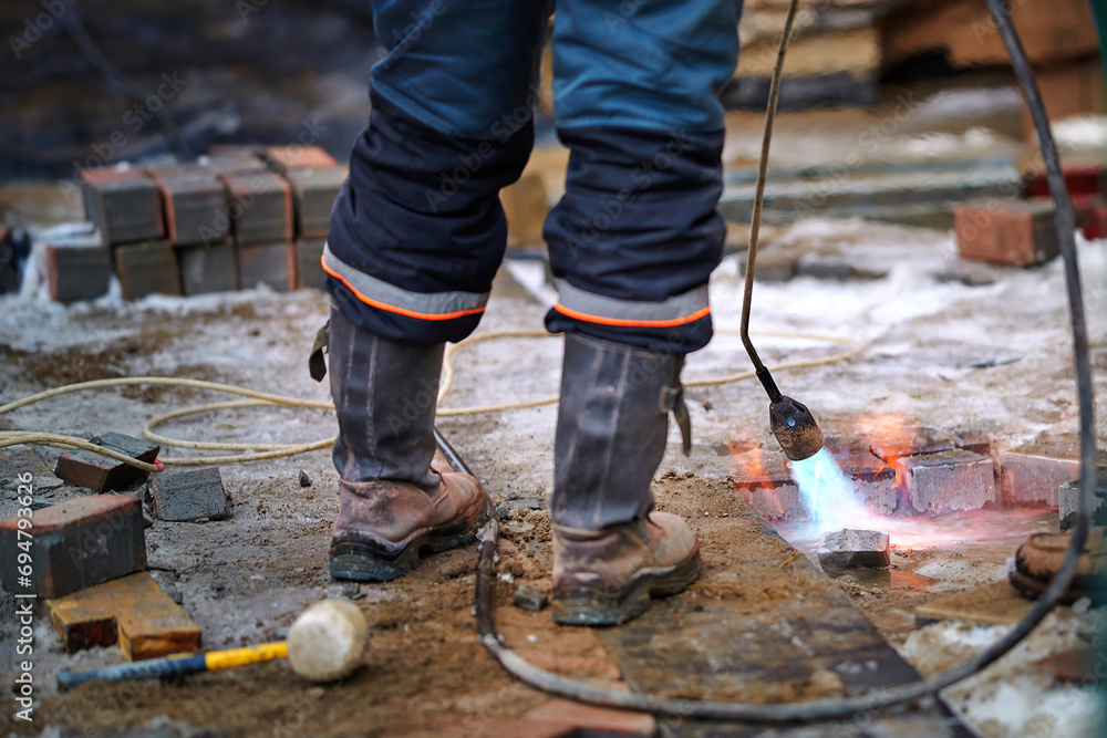 Worker dries wet surface with gas torch before paving work in winter season. Laying concrete paving slabs in cold season outdoors. Bricklayer working. Repair, restoration of sidewalks. Selective focus Worker dries wet surface with gas torch before paving work in winter season. Laying concrete paving slabs in cold season outdoors. Bricklayer working. Repair, restoration of sidewalks. Selective focus