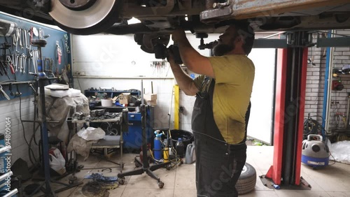 Auto mechanic working underneath a lifting vehicle at garage. Professional bearded repairman repairing car with special electrical tool at workshop. Concept of automobile maintenance. Slow motion photo