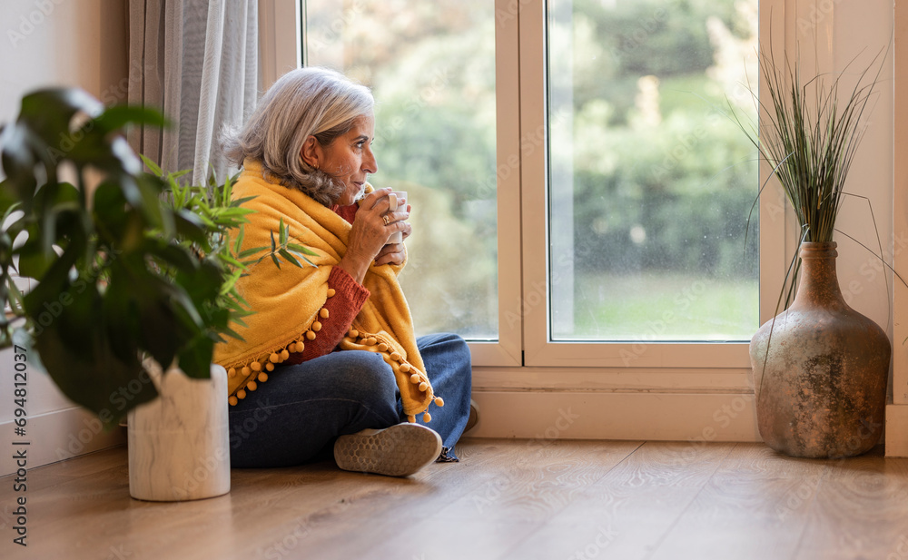 senior woman sitting on the floor of her apartment drinking hot cup drink next to the window covered senior woman sitting on the floor of her apartment drinking hot cup drink next to the window covered