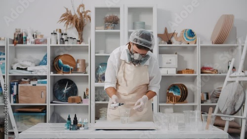 Female artist pouring paint into cup with art epoxy resin working in art studio workshop, creative woman in protective face mask engaged in trendy hobby creating beautiful sea waves acrylic paintings photo