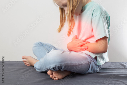 A seven-year-old blonde girl sits and holds her red belly. Concept of pain and cramps in the abdomen. Stomach diseases, gastritis and peptic ulcers in children. Copy space for text, dysfunction photo