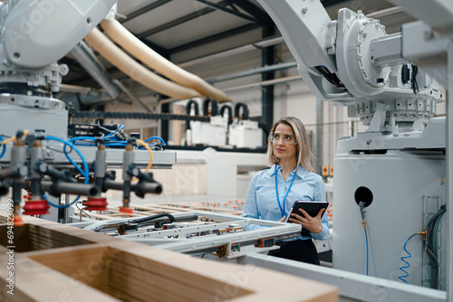Female engineer in modern industrial factory, checking robotic arm, controling process and manufacturing equipment or machinery. Manufacturing facility with robotics and automation.