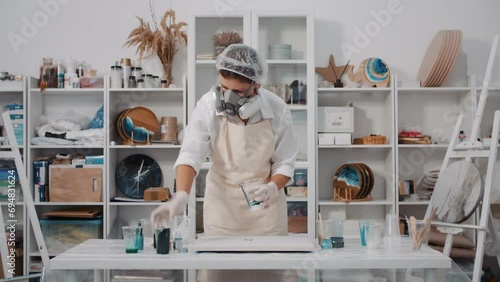 Female artist mixing colored art epoxy resin in plastic cup working in art studio workshop, creative woman in protective mask engaged in trendy hobby creating beautiful sea waves acrylic paintings photo