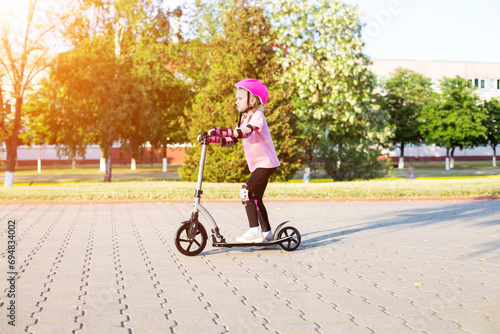 A beautiful girl of 7 years old rides a scooter in a pink protection against injuries and falls. Sunny summer day. Copy space for text