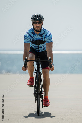 Front view of fit young man cycling in the beach looking at camera. Athletic cylcist training and riding a bike wearing helmet and sunglasses with sea in the background.