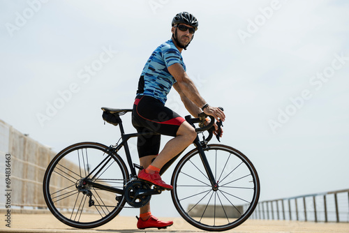 Side view of a fit man standing on bike wearing helmet and sunglasses. Athletic male cyclist training with cycle outdside looking at camera.