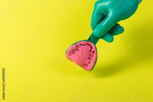 The hand of a dentist doctor in a green medical glove holds a dental spoon with an alginate plastic mass for making a plaster cast of the patient s jaw on a yellow background. Concept of malocclusion photo