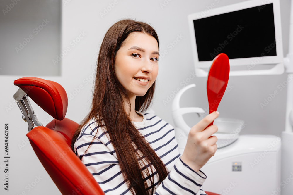 Happy caucasian female patient looking at mirror after dental treatment in clinic. Cheerful woman sitting in chair in dental cabinet and enjoying her beautiful smile Happy caucasian female patient looking at mirror after dental treatment in clinic. Cheerful woman sitting in chair in dental cabinet and enjoying her beautiful smile
