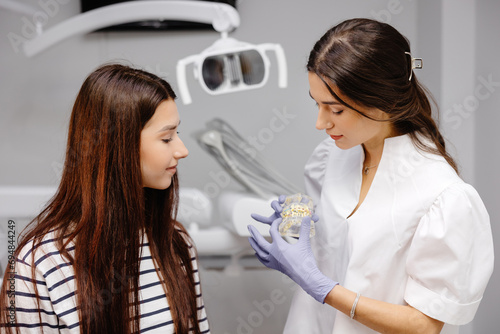 Young woman having a visit at the dentist. Female doctor showing fake plastic jaw. Beautiful girl patient sitting on chair at dentist office in dental clinic. Healthy teeth and medicine concept.