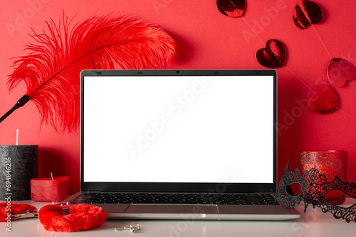 Explore virtual desires with side view of laptop, candles, mask, and adult toys—furry handcuffs and feather tickler on red background. An empty screen invites you to embark on a sensual online journey