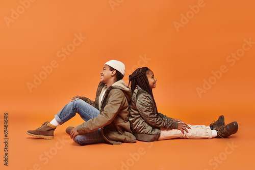 preteen african americna kids in warm outfits sitting on floor back to back on orange backdrop photo