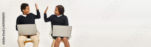 happy african american schoolkids in uniform using laptops and giving high five on grey, banner