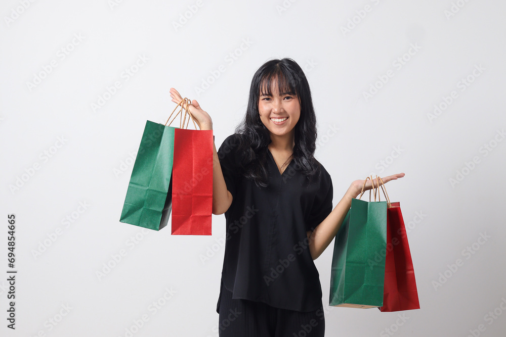 Portrait of excited Asian woman in black shirt holding and showing colorful paper shopping bags. Shopaholic girl and discount buying concept. Isolated image on white background