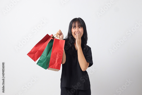 Portrait of excited Asian woman in black shirt holding and showing colorful paper shopping bags. Shopaholic girl and discount buying concept. Isolated image on white background