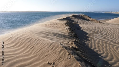 Heavy wind blows sand over the ridge of a sand dune towards the sea. Lac Naila, where the Sahara desert meets the Atlantic Ocean, National Park Kheniffis, Morocco. 4k natural landscape background.