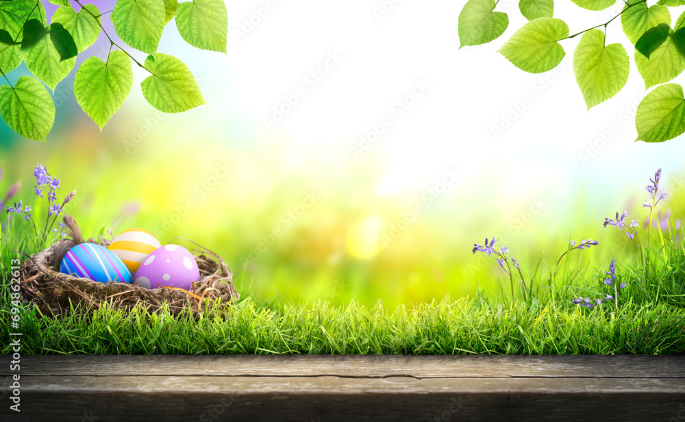 Three painted easter eggs in a birds nest celebrating a Happy Easter in spring with a green grass meadow, tree leaves and bright sunlight background with copy space & wooden bench to display products.