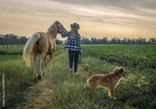 young blonde girl in a hat and a plaid shirt walks with a horse and dog on a farm in the village