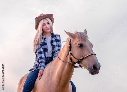 young girl dressed in a cowboy hat and blue jeans sits on a beige horse on the ranch © Sofiia