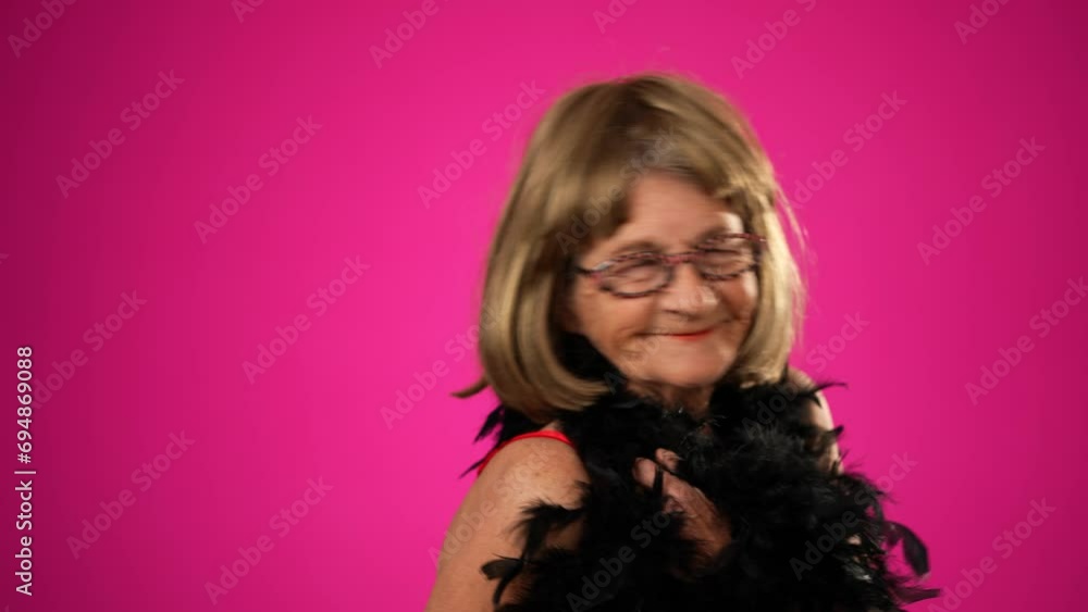 Funny elderly woman walks across frame throws boa over shoulder, and smiles at camera. Portrait of old woman isolated on pink background Funny elderly woman walks across frame throws boa over shoulder, and smiles at camera. Portrait of old woman isolated on pink background