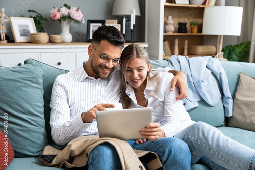 Excited young couple sitting on couch in formal wear with tablet pc, celebrating online win, great deal or business success at home. Millennial spouses enjoying big sale in web store. photo