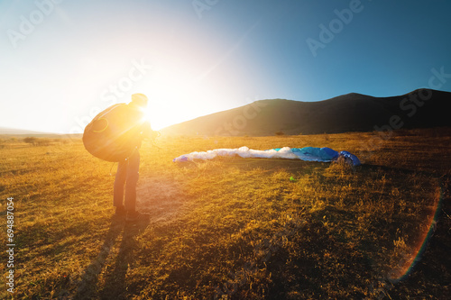 A paraglider takes off from a mountainside with a blue and white canopy and the sun behind. A paraglider is a silhouette. The glider is sharp, with little wing movement.