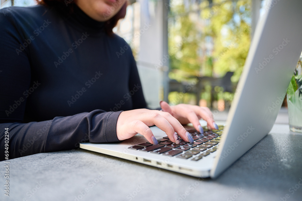Naklejka premium Cropped image of the student girl typing on laptop while sitting at a table in a cafe. Female student studying at park using modern laptop at bright sunny day. Working outdoor.