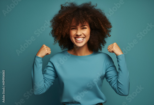Waist up shot of cheerful curly haired woman keeps hand behind head smiles toothily enjoys happy moment in life wears big spectacles purple turtleneck isolated over blue background. Positive emotions