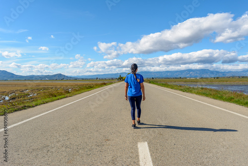 A person is walking down an open road surrounded by nature under a cloud-streaked blue sky  back view of latina woman dressed in blue walking down the road in the Ebro Delta natural park  Tarragona