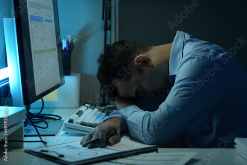 Young Asian businessman experiences stress, headaches, eye strain and fatigue while working with head down on PC keyboard and stack of financial documents on table. Office Syndrome.